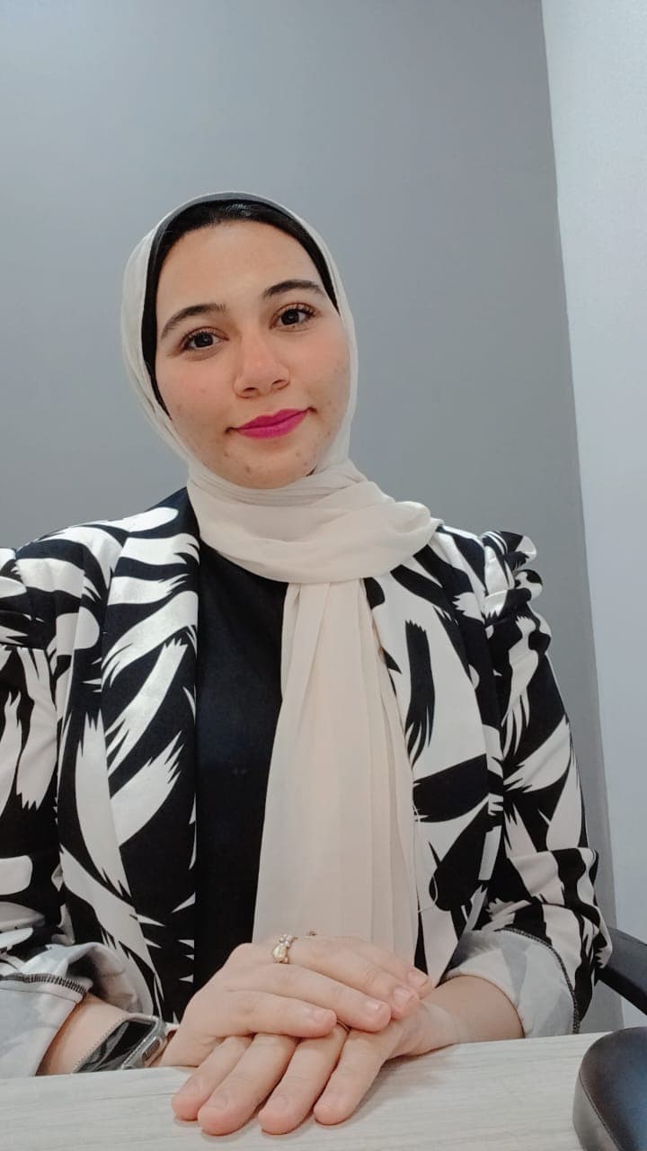 امرأة مبتسمة ترتدي حجاباً أبيض وسترة بنقوش سوداء وبيضاء تجلس خلف مكتب عمل.